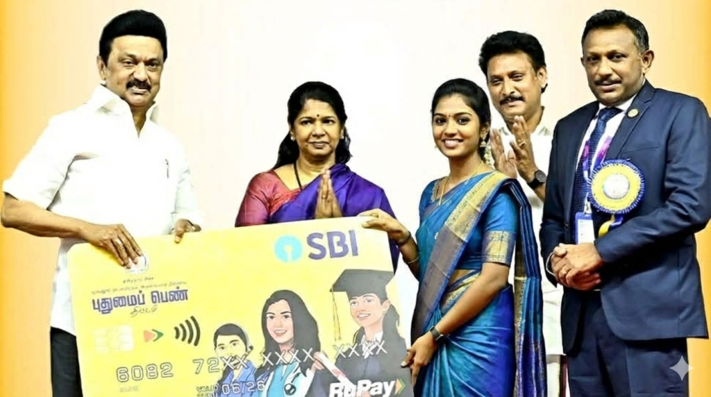 A photograph showing a group of officials, including Tamil Nadu Chief Minister M.K. Stalin, presenting a large ceremonial SBI "Pudhumai Penn" RuPay card to a smiling 20-year-old Tamil woman. The woman is dressed in a traditional royal blue silk saree with gold borders, gold jewelry, and jasmine flowers in her hair. She is flanked by other dignitaries against a plain, light background.