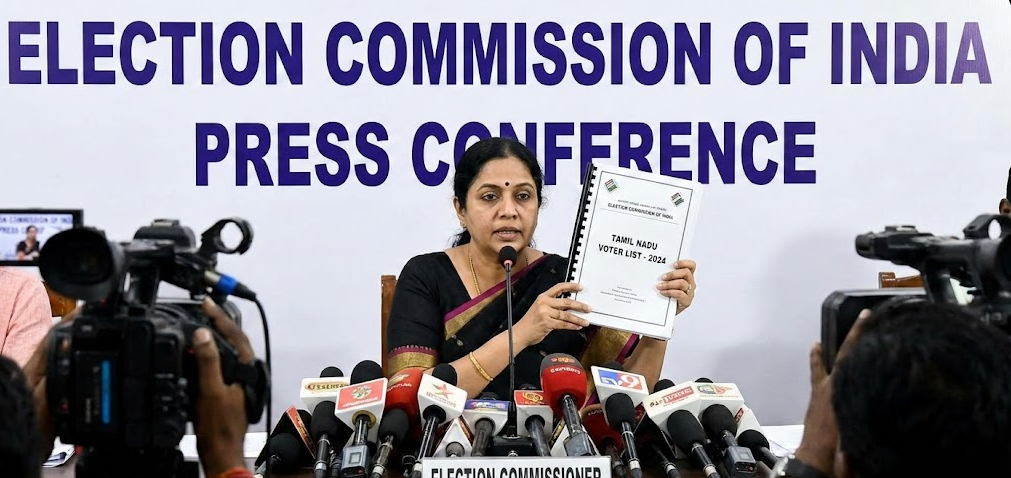 A professional female election commissioner seated at a press conference table, holding up a bound document titled "TAMIL NADU VOTER LIST - 2024." Behind her is a large banner for the "Election Commission of India," and several news microphones and cameras are positioned in the foreground to capture the announcement.